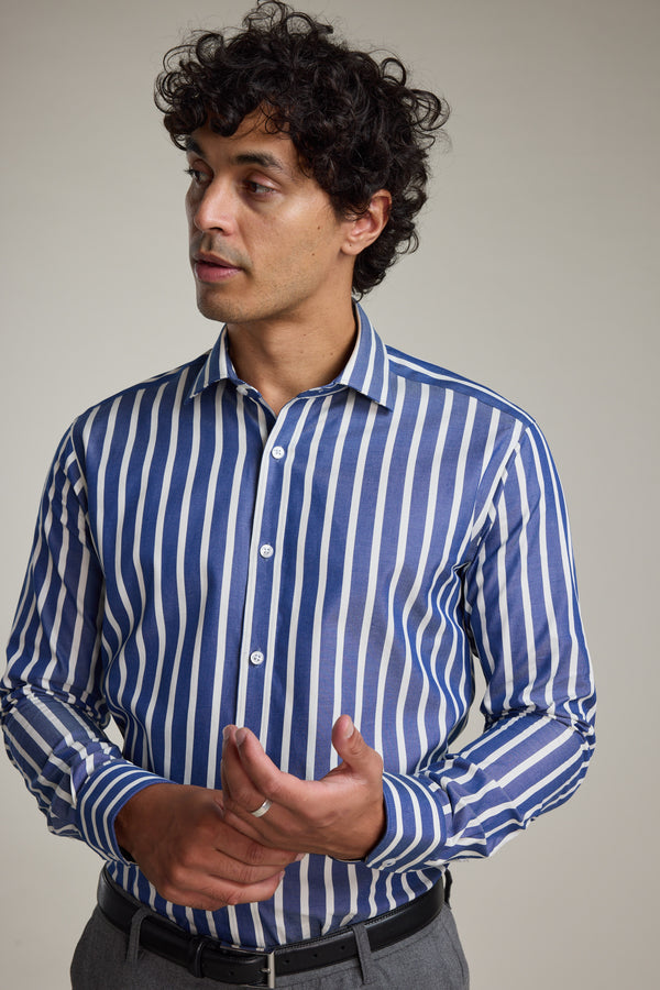 A man with curly dark hair wears the Barkers Nandas Stripe Business Shirt—blue and white vertical stripes—and gray pants, standing against a plain background, looking left with his hands clasped together.