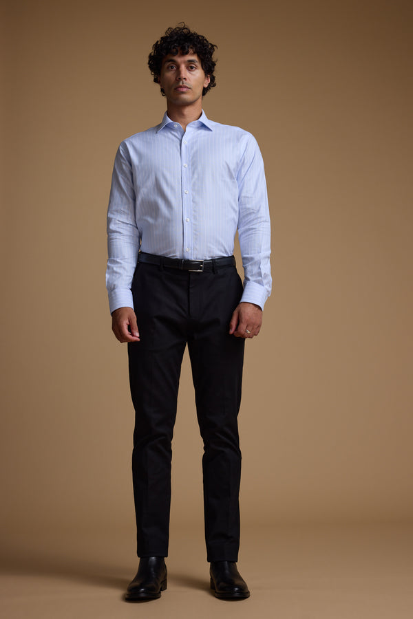 A man with curly hair stands facing forward against a plain brown background, wearing Barkers’ Goodall Stripe Business Shirt with black pants, a black belt, and black shoes.