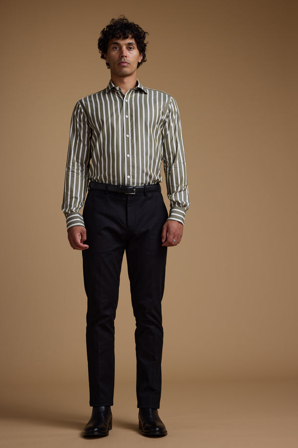 A man with curly hair stands against a plain brown background, wearing the Barkers Nandas Stripe Business Shirt—a wrinkle-resistant, long-sleeve green and white shirt—paired with dark trousers and black shoes.