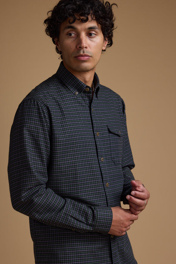 A man with curly brown hair wears the Barkers Starle Check Shirt, a vintage-style design made from responsibly sourced cotton, standing against a plain beige background and looking slightly to his left with a neutral expression.