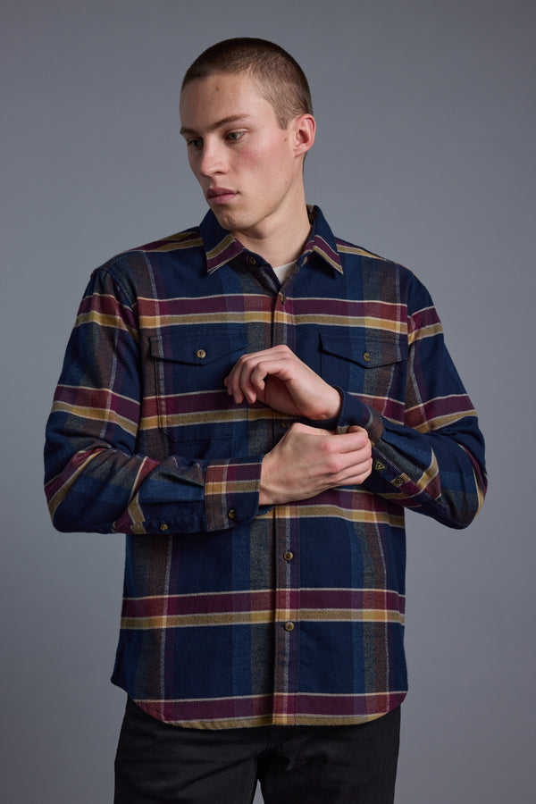 A young man in the Barkers Heaton Check Overshirt, crafted from responsibly sourced cotton, stands against a plain gray background and looks left while adjusting his sleeve.