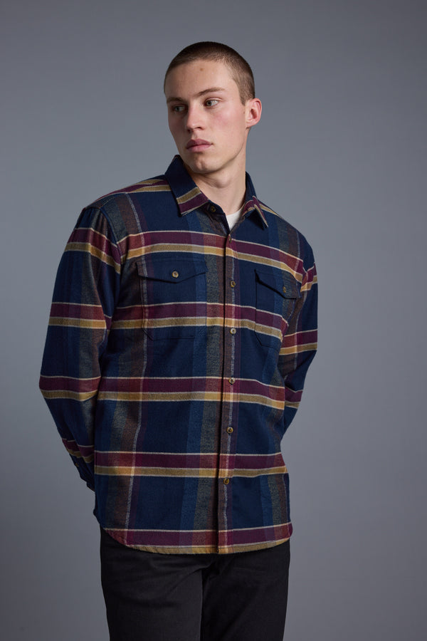 A young man stands against a gray background, hands behind his back and looking to his right, wearing the Barkers Heaton Check Overshirt—a vintage plaid flannel shirt in blue, maroon, and yellow made from responsibly sourced cotton.