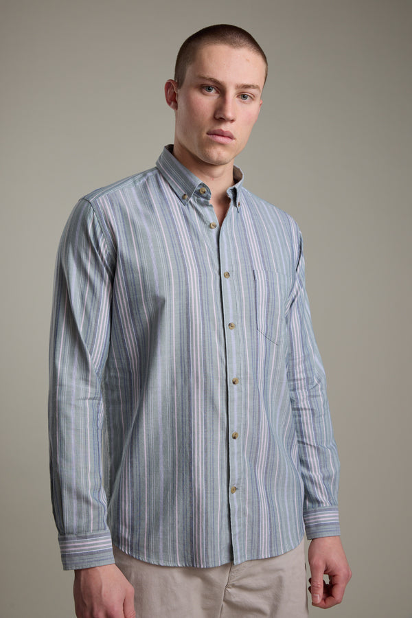 A young man with a buzz cut wears the Barkers Huntley Striped Oxford Shirt, featuring vertical pastel stripes and tailored fit, paired with beige pants against a plain light gray background.