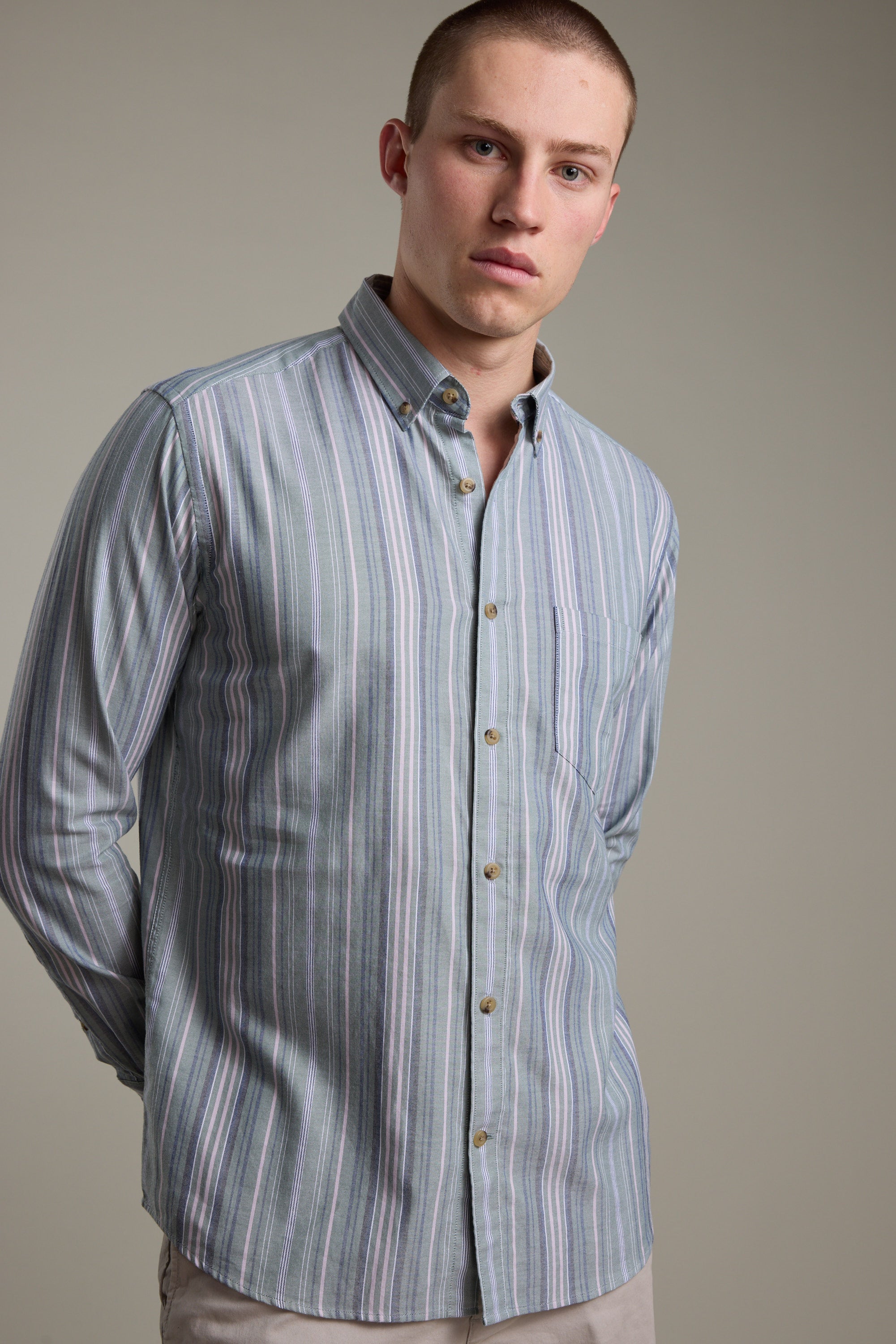 A young man with short hair models the Barkers Huntley Striped Oxford Shirt, featuring vertical blue, white, and pink stripes in breathable cotton. He stands against a plain background, hands behind his back, gazing at the camera.