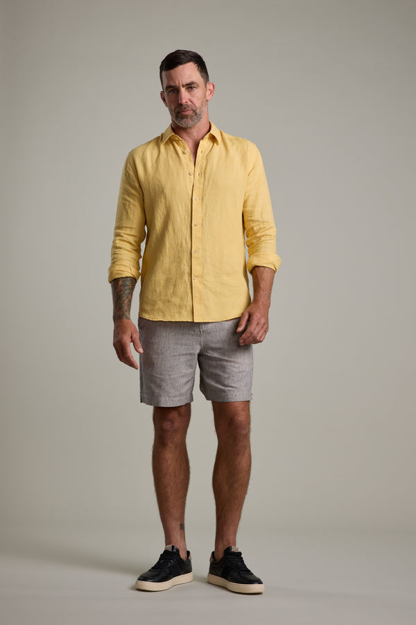 A man with short hair and a trimmed beard stands confidently against a plain background wearing the Barkers Dorset Linen Shirt in light yellow, paired with gray striped shorts and black sneakers.