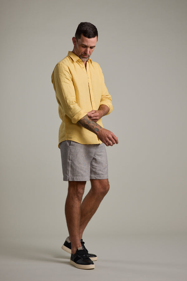 A man adjusts his sleeve, wearing the Barkers Dorset Linen Shirt in yellow, paired with gray striped shorts and black sneakers against a plain, light-colored background.