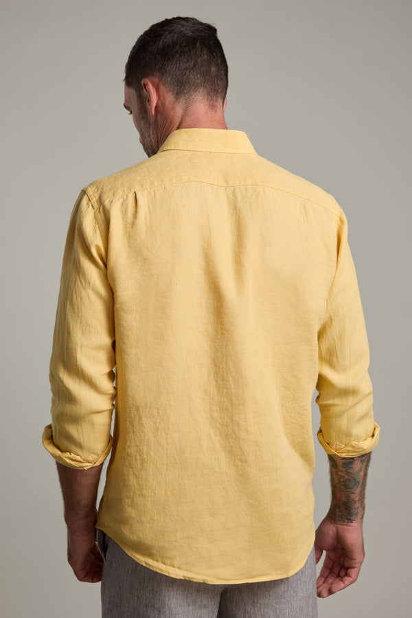 A man with short hair, tattooed left forearm, and light pants stands facing away in a Barkers Dorset Linen Shirt—tailored fit, long-sleeved, light yellow French linen, sleeves rolled up.
