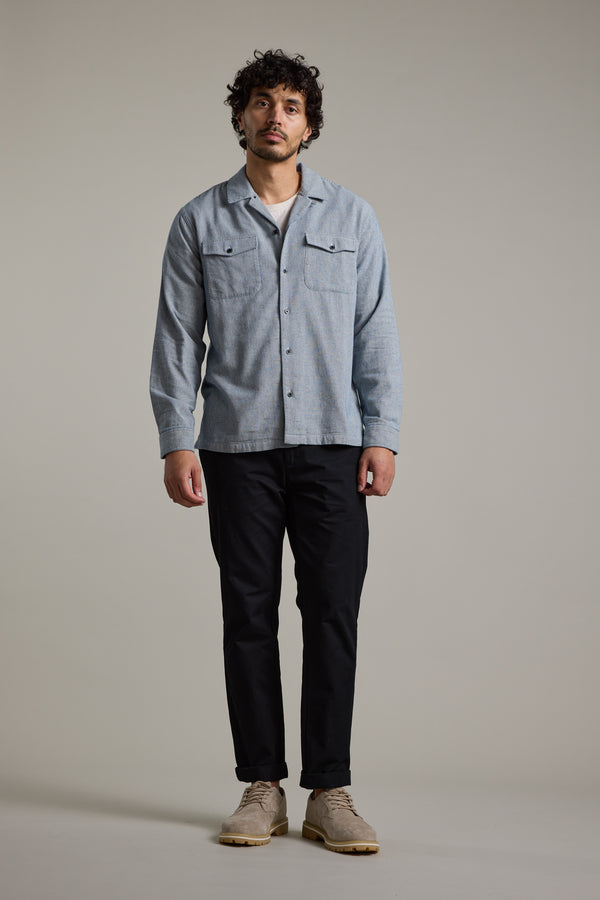 A man with curly hair stands facing forward against a plain background, wearing the Barkers Sherman Cuban Overshirt in light blue cotton over a white t-shirt, paired with black pants and beige shoes.