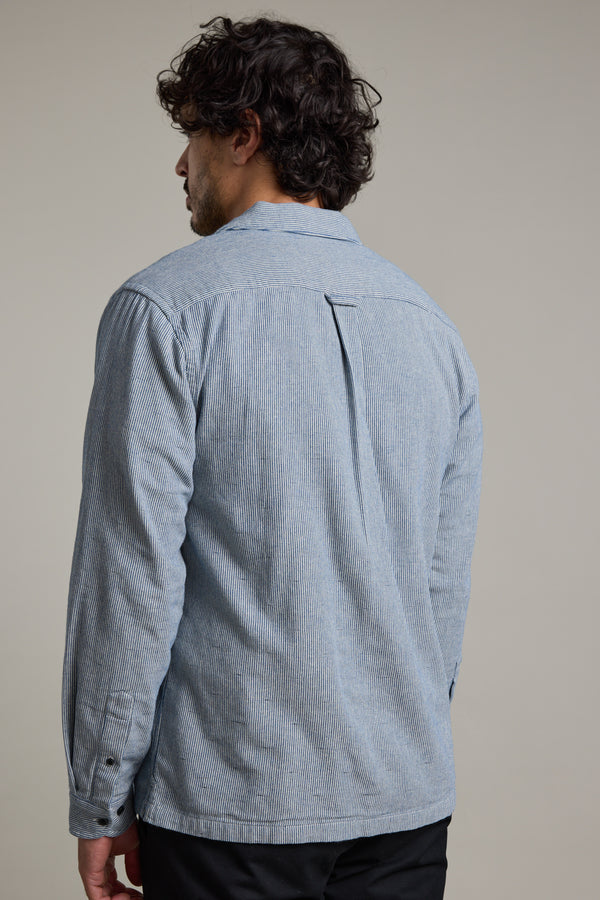 A man with curly dark hair stands with his back to the camera, wearing Barkers' Sherman Cuban Overshirt in a relaxed fit light blue with subtle vertical stripes, paired with black pants against a plain background.