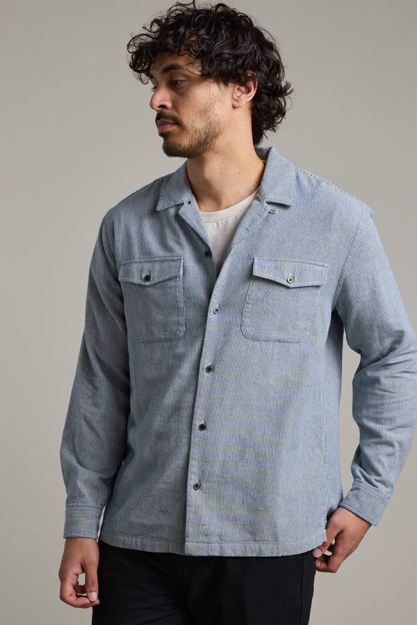 A man with curly hair and a beard wears the Barkers Sherman Cuban Overshirt in light blue over a white T-shirt, standing against a plain background and looking left with his right hand in his pocket.