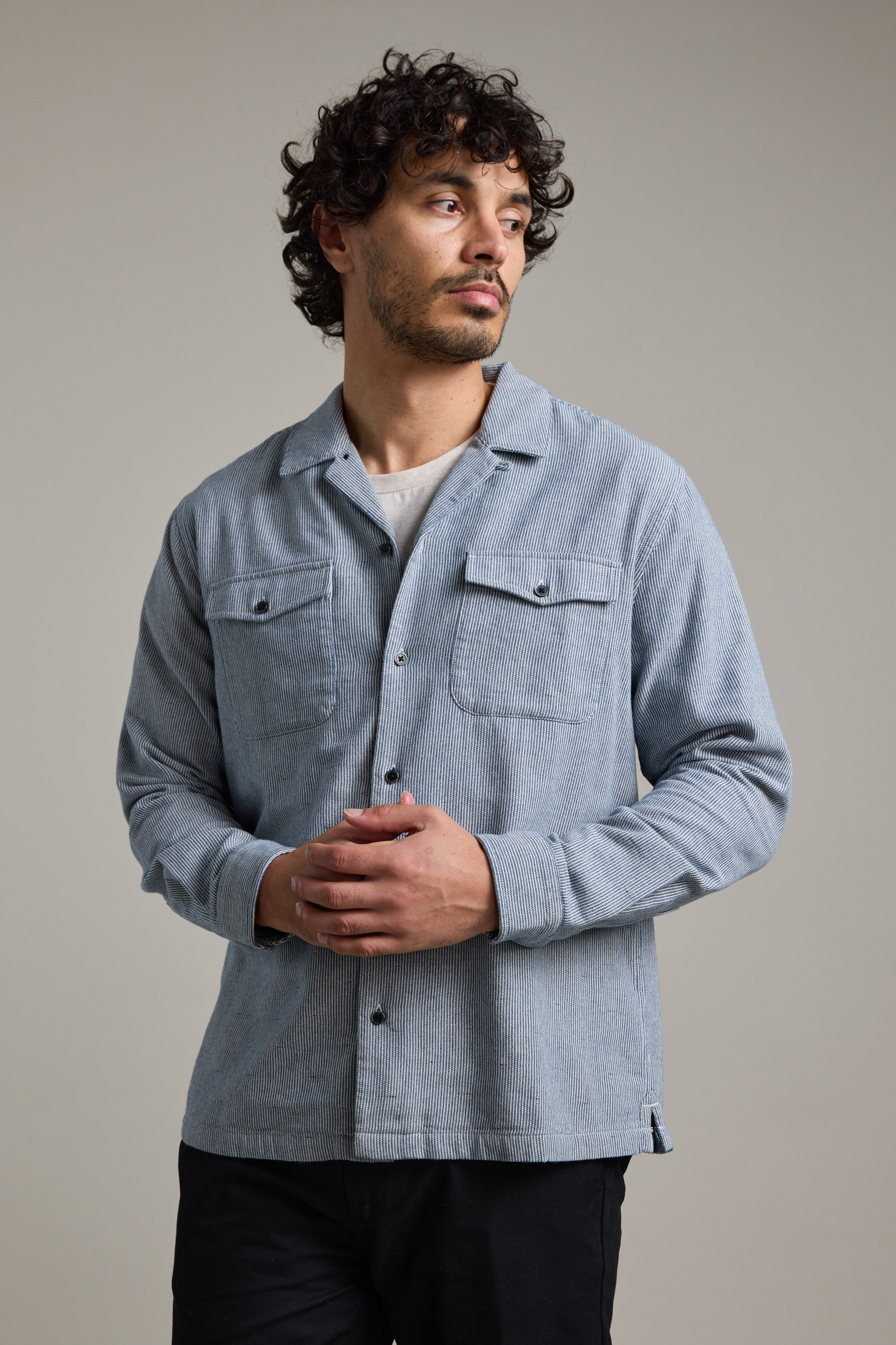 A man with curly hair and a beard wears the Barkers Sherman Cuban Overshirt in light blue over a beige t-shirt, standing with hands clasped and looking to his left against a plain neutral background.