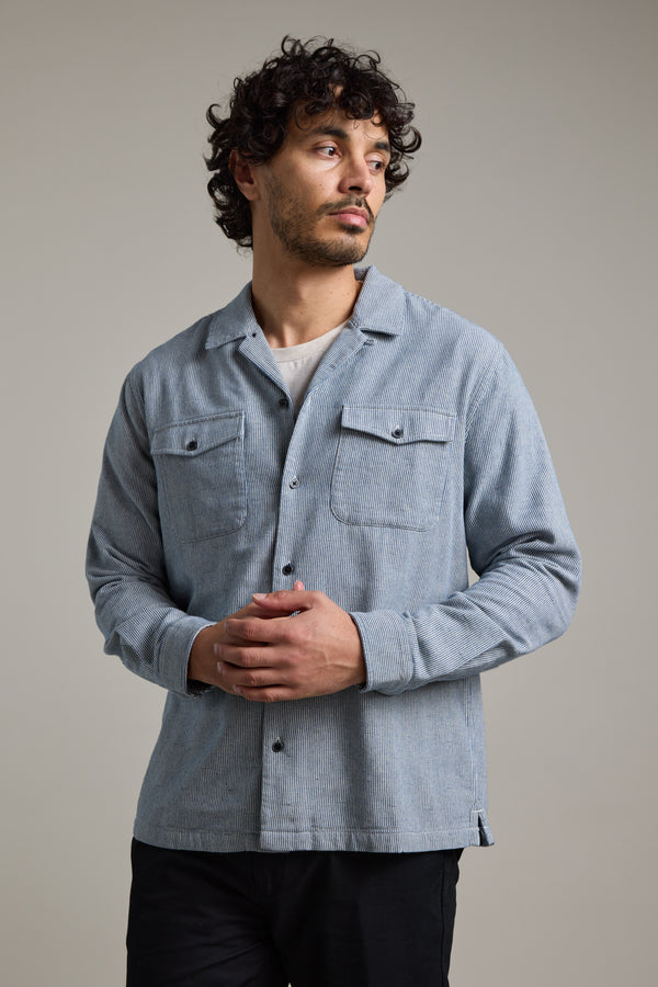 A man with curly hair and a beard wears the Barkers Sherman Cuban Overshirt in light blue over a beige t-shirt, standing with hands clasped and looking to his left against a plain neutral background.