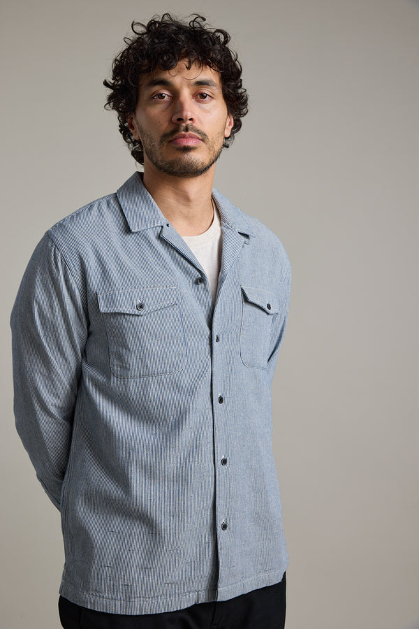 A man with curly hair and a short beard stands confidently against a plain background, wearing the Barkers Sherman Cuban Overshirt in light blue textured cotton over a white t-shirt, with his hands behind his back.