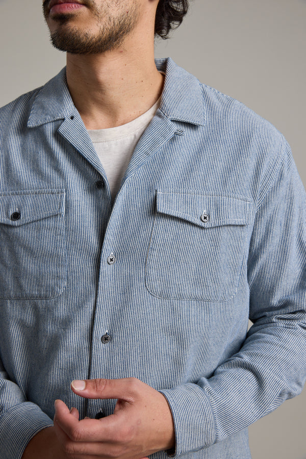 A man wears the Barkers Sherman Cuban Overshirt, a relaxed fit blue and white striped shirt, over a white T-shirt. He stands with hands loosely clasped, face partially visible against a plain neutral background.