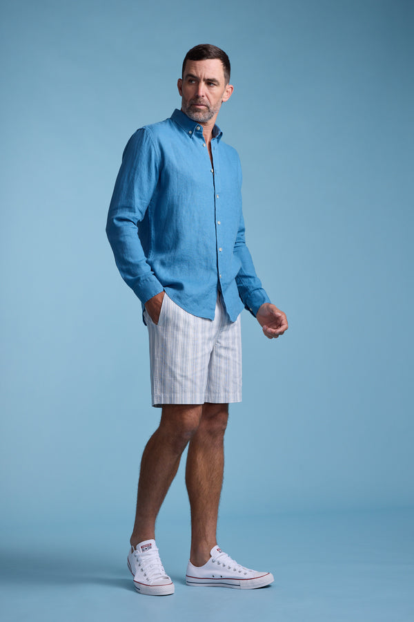A man stands against a light blue background wearing the Barkers Dorset Linen Shirt, light-colored striped shorts, and white sneakers. He gazes slightly to the side with one hand in his pocket.