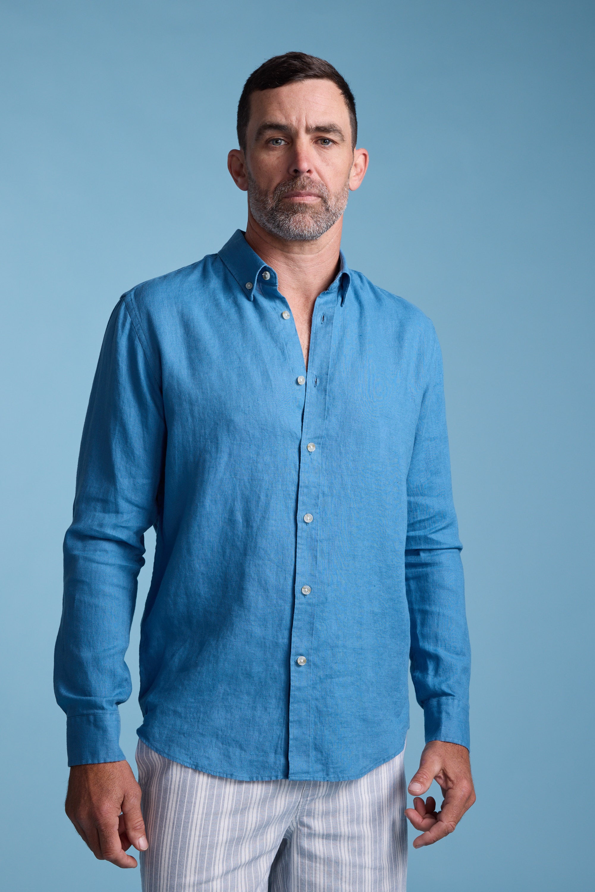 A man with short dark hair and a beard wears the Barkers Dorset Linen Shirt and light-colored striped pants, standing against a plain light blue background.