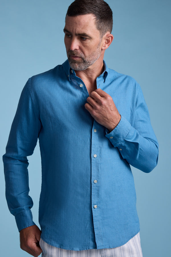 A man with short dark hair and a trimmed beard wears the Barkers Dorset Linen Shirt with light striped pants, standing against a light blue background while adjusting his shirt collar.
