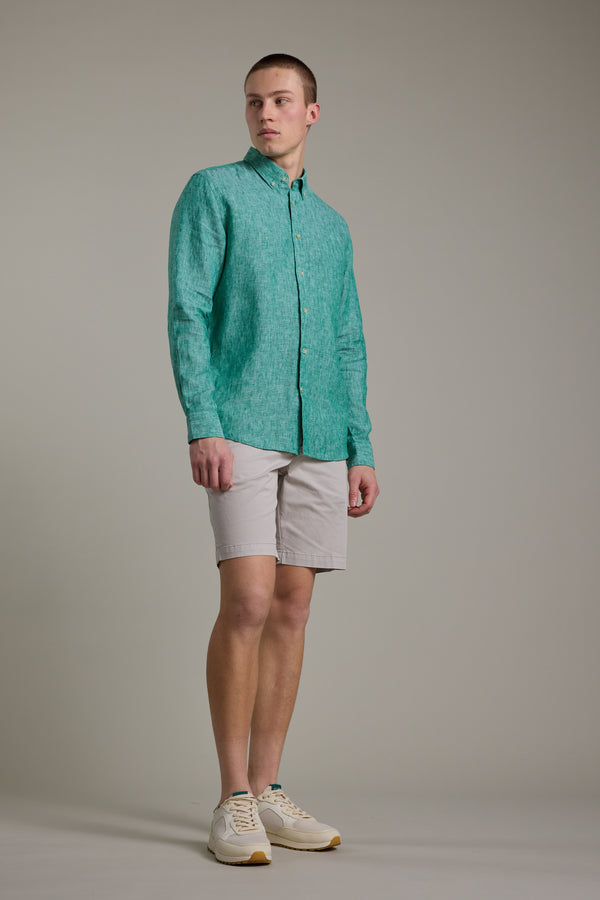 A young man stands against a plain background wearing the Barkers Dorset Linen Shirt in teal, paired with light beige shorts and white sneakers. He looks slightly to his left with a neutral expression.