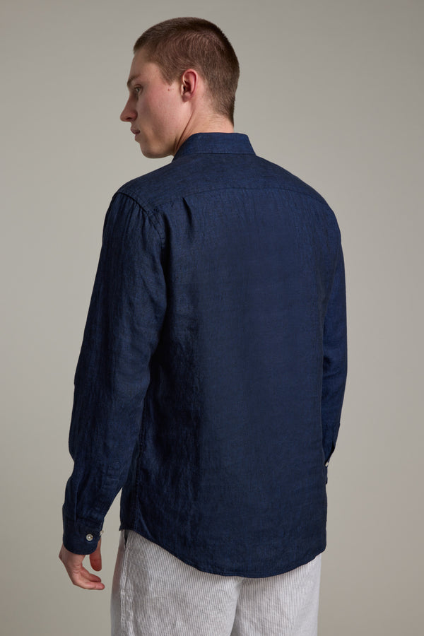 A person with short hair is seen from behind, dressed in a Barkers Dorset Linen Shirt in dark blue and light-colored pants, standing against a plain, light background.