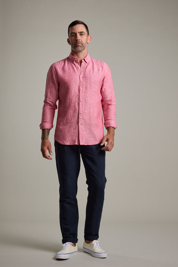A man with short hair and a trimmed beard stands against a plain background wearing the Barkers Dorset Linen Shirt in pink, styled with dark pants and white sneakers. He looks straight ahead with a neutral expression.