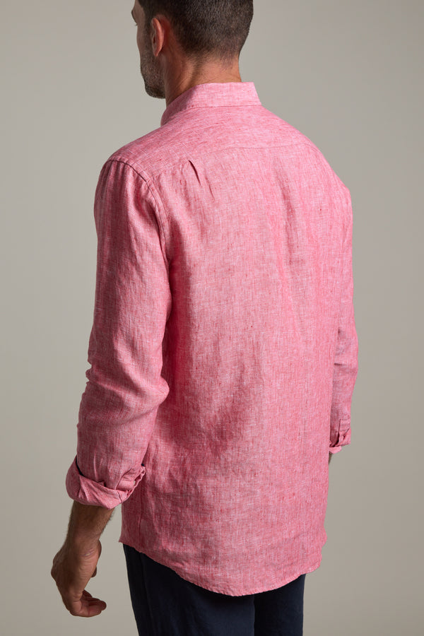 A man, seen from behind, wears the Barkers Dorset Linen Shirt in long-sleeve light red French linen with rolled-up sleeves and dark pants, standing against a plain, neutral background.