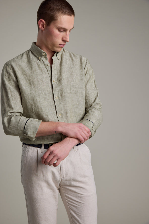 A man wears the Barkers Dorset Linen Shirt in light green with rolled-up sleeves and light beige pants, adjusting his sleeve against a plain background to highlight the shirt's breathable linen and tailored fit.