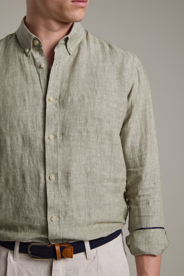 A man stands against a plain background wearing the Barkers Dorset Linen Shirt in light olive green, featuring rolled-up sleeves, paired with beige pants and a navy woven belt with a tan leather buckle.
