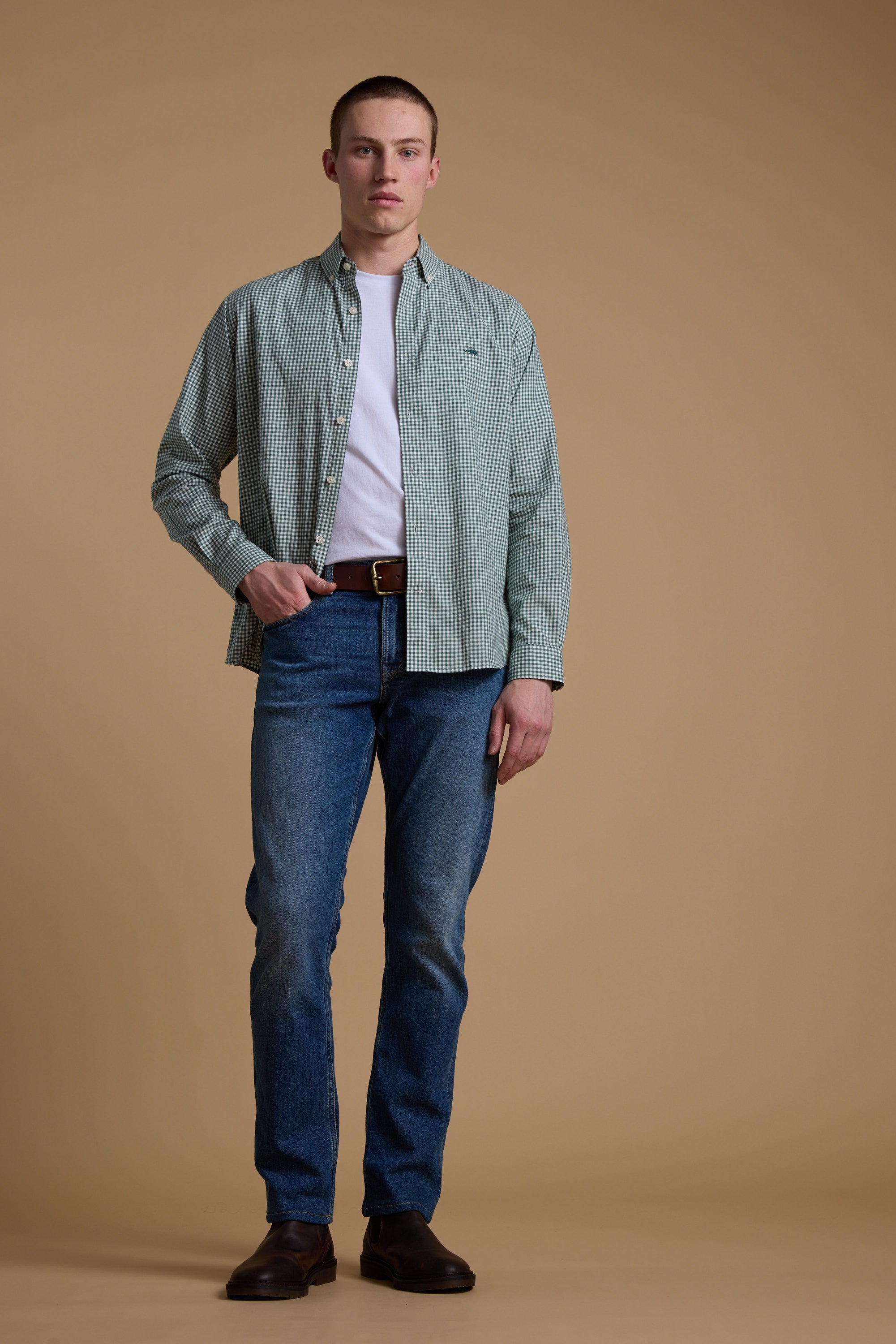 A man stands against a plain beige background wearing a green Barkers Courtland Check Shirt over a white tee, blue jeans, brown belt, and dark brown shoes. He looks forward neutrally. The shirt is made from responsibly sourced cotton.