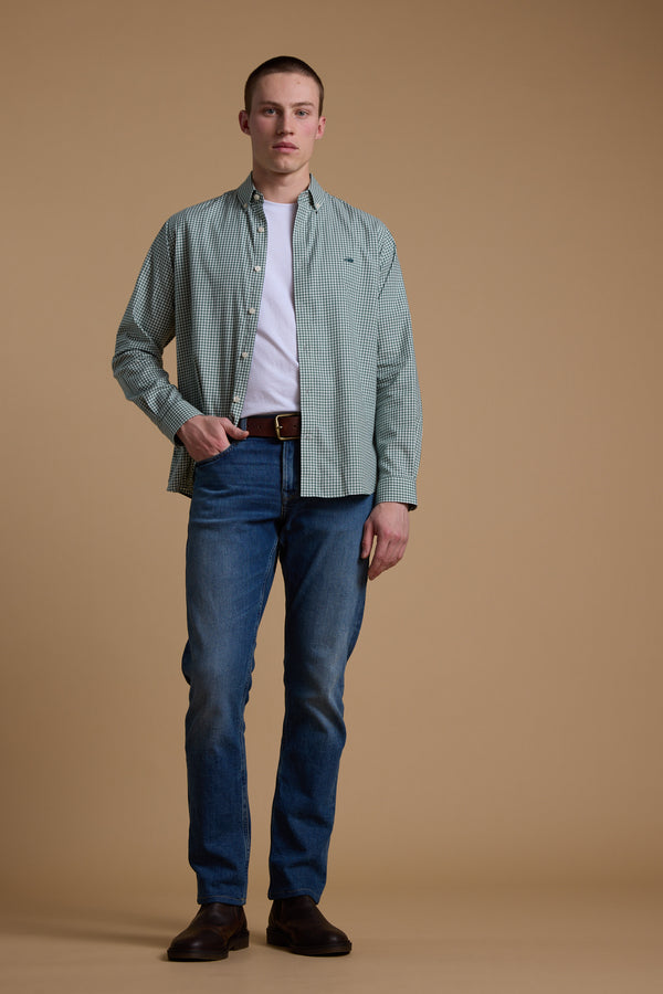A man stands against a plain beige background wearing a green Barkers Courtland Check Shirt over a white tee, blue jeans, brown belt, and dark brown shoes. He looks forward neutrally. The shirt is made from responsibly sourced cotton.