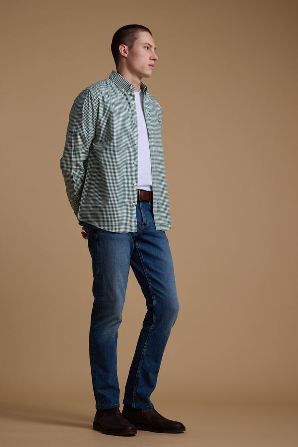 A man stands against a plain beige background, wearing a green checkered Barkers Courtland Check Shirt over a white t-shirt, paired with blue jeans, a brown belt, and brown shoes. He looks to the side with his hands behind his back.