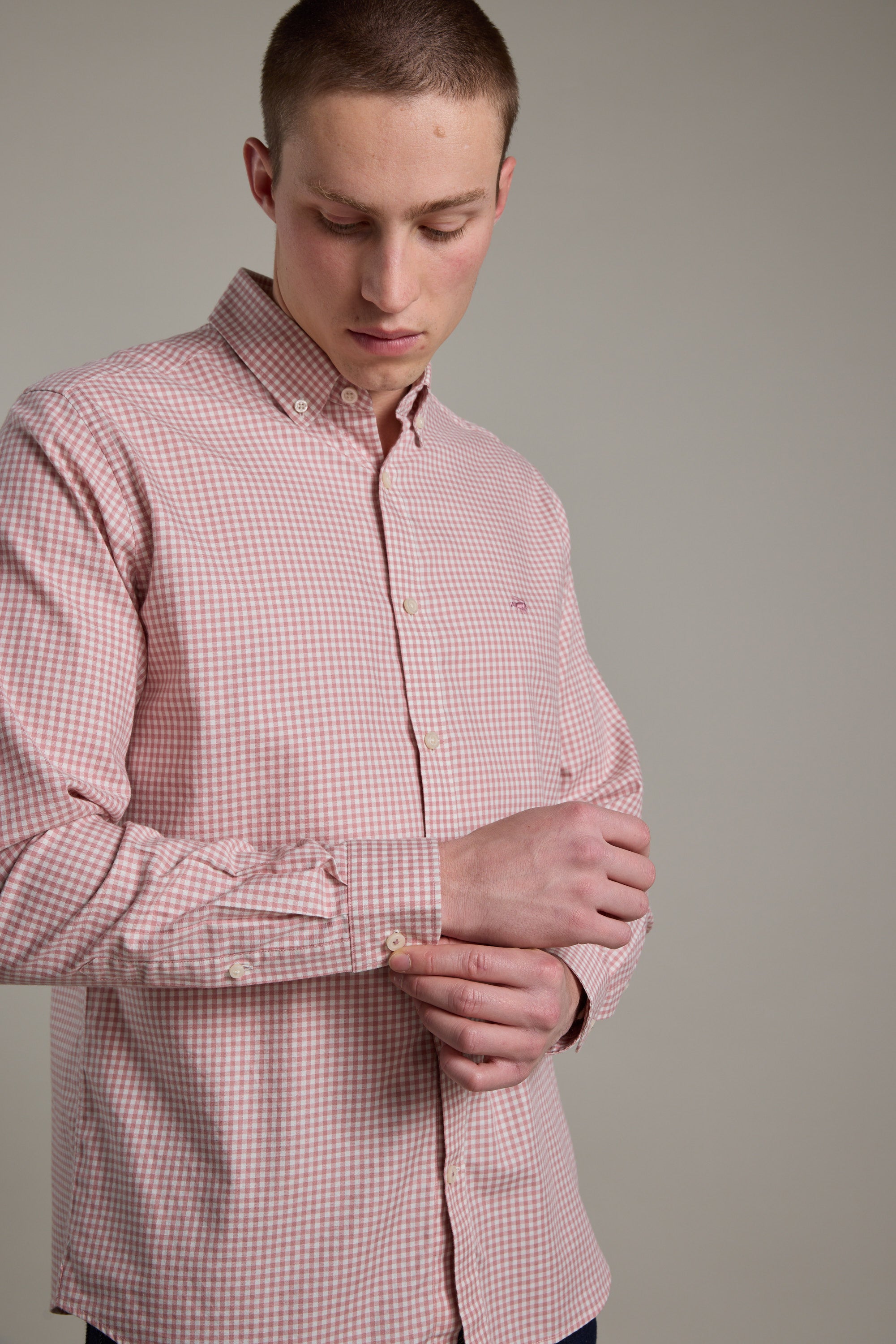 A man adjusts his left cuff while wearing the Barkers Courtland Check Shirt, made from responsibly sourced cotton in a light pink and white check pattern, against a plain, light background.