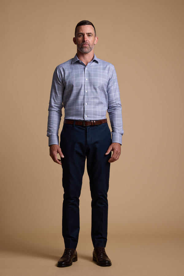 A man with short dark hair and a beard stands facing forward against a plain tan background, wearing the Barkers Kenmore Check Business Shirt in light blue, paired with tailored dark blue trousers, a brown belt, and brown dress shoes.