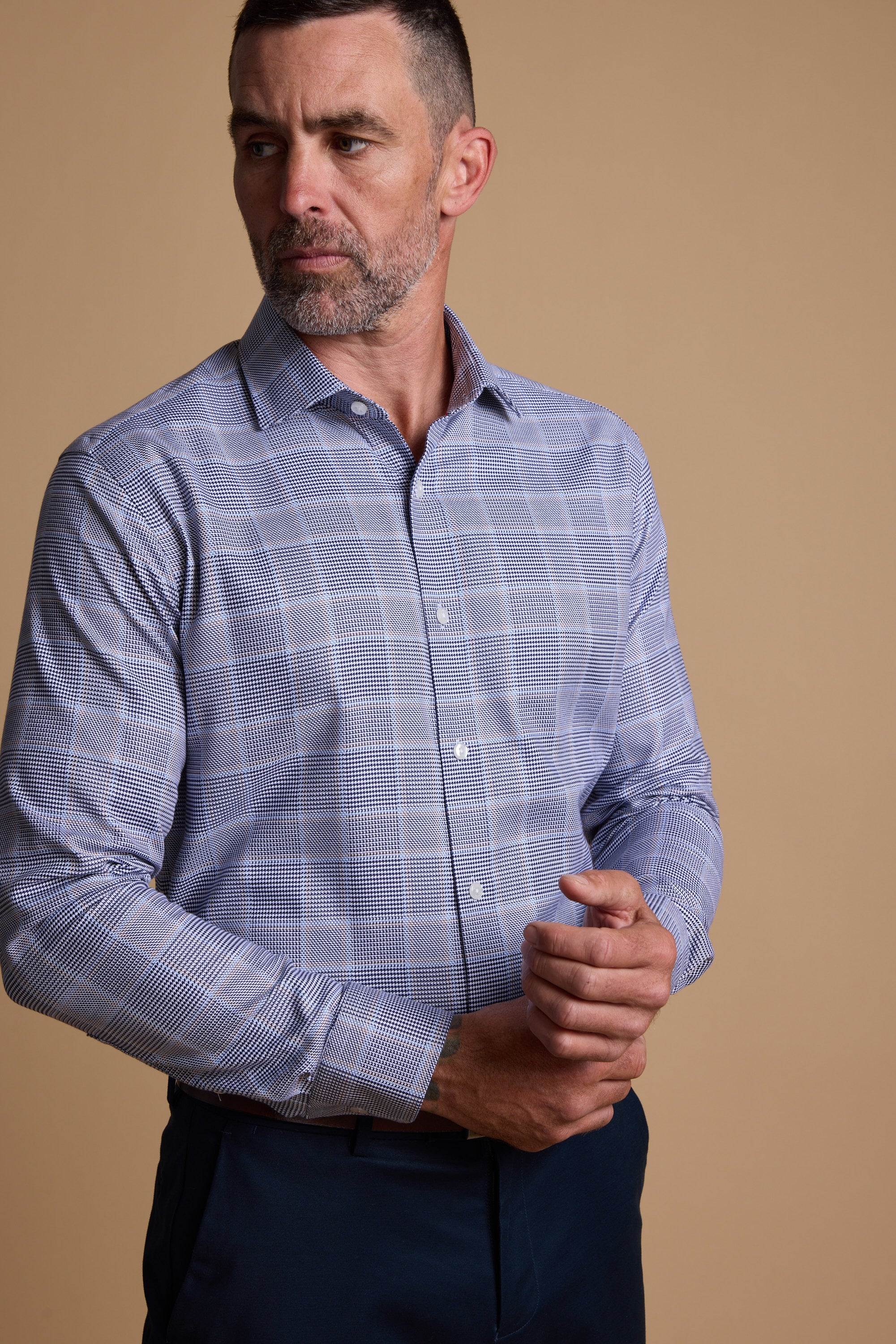A man with short dark hair and a beard wears the Barkers Kenmore Check Business Shirt, a tailored fit, wrinkle-resistant shirt in light blue and white checks, standing against a beige background while adjusting his cuff.