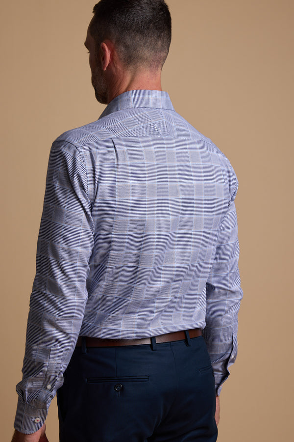 A man with short hair is shown from the back wearing the Barkers Kenmore Check Business Shirt, a wrinkle-resistant, light blue plaid shirt, and dark blue trousers against a plain beige background.