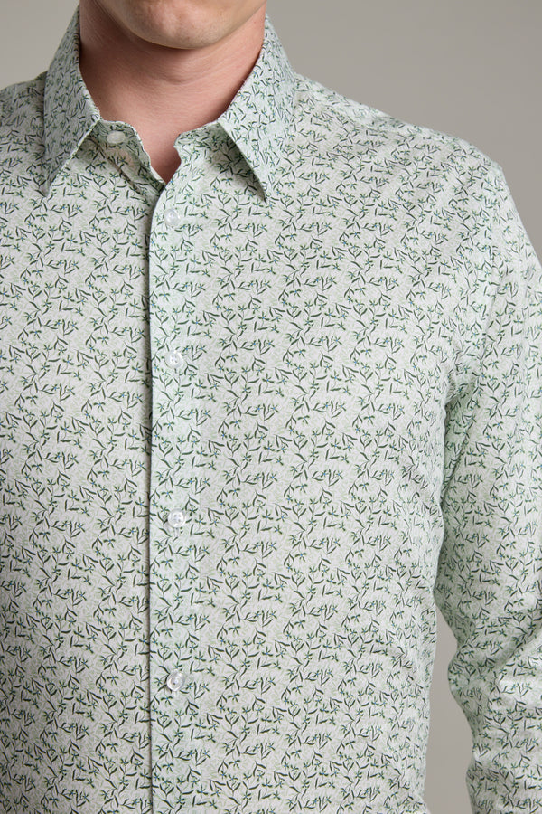 A person models the Barkers Ambrose Print Shirt, featuring a light green base with a small dark green and black leafy pattern, made from responsibly sourced cotton, shown chest up against a plain background.