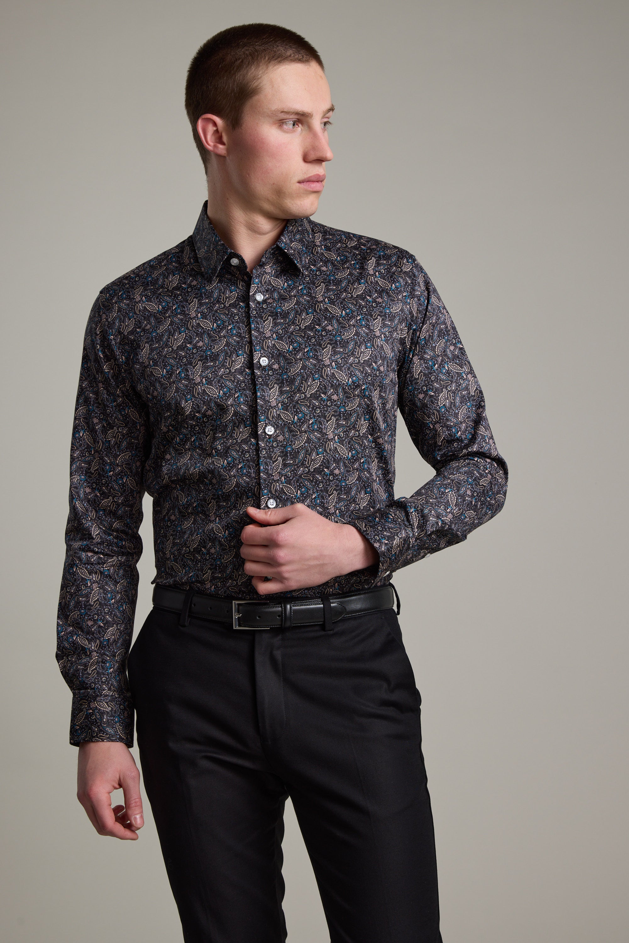 A man in a tailored-fit Barkers Hilton Print Shirt with a subtle floral pattern and black trousers stands against a light gray background, looking left with one hand near his buttons, showcasing a modern, stylish appearance.
