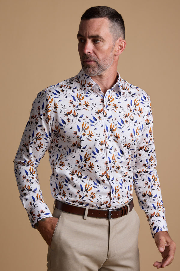 A man with short dark hair and a beard wears the Barkers Milo Print Shirt, a white shirt with a colorful floral pattern, paired with beige trousers. He stands against a tan background, looking left with one hand in his pocket.