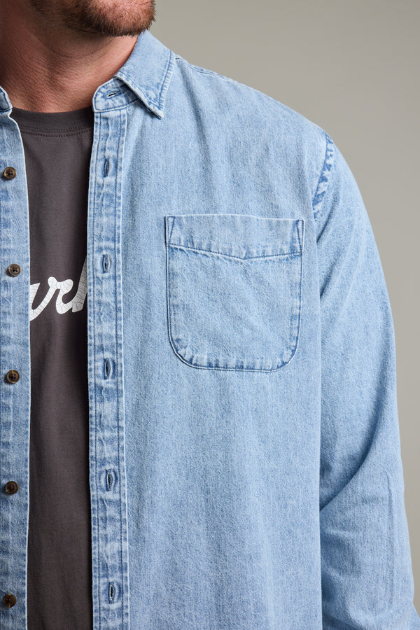 A man models the Barkers Ruxton Denim Shirt, a classic-fit light blue overshirt, layered over a dark tee with white lettering; only his face’s lower half, chest, and upper arm are visible.