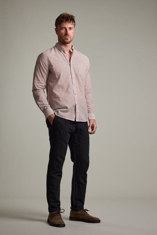 A man stands against a plain background wearing the Barkers Sumter Stripe Shirt—a lightweight, light pink and white striped tailored fit—paired with dark pants and brown shoes, hands in pockets and a neutral expression.