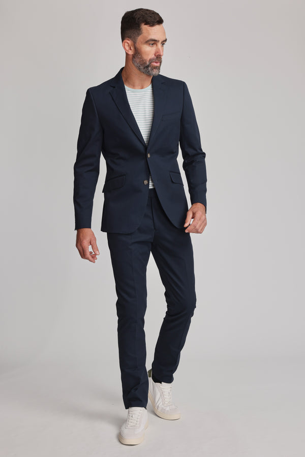 A man with short dark hair and a beard models the Barkers Maclay Twill Blazer over a light striped tee and white sneakers, standing against a plain light background and looking to the side.