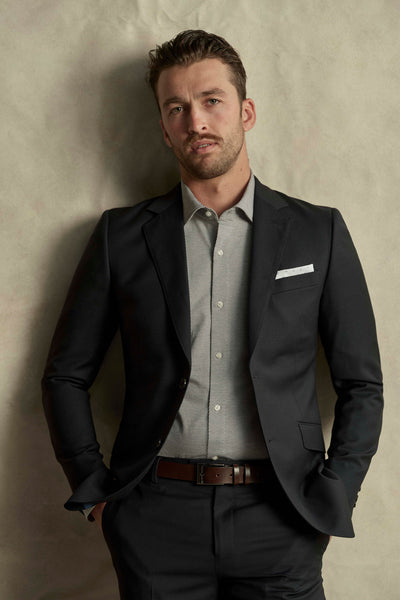 A man stands against a textured beige background, hands in pockets, wearing a Barkers Maclay Twill Blazer in black over a light gray dress shirt, looking at the camera with a neutral expression.