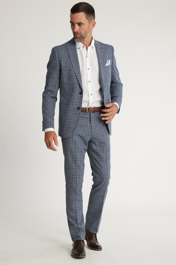 A man in Barkers Highland Check Suit Pant, a blue plaid suit, white dress shirt, brown belt, and brown dress shoes stands against a plain white background, looking down and slightly to his left.