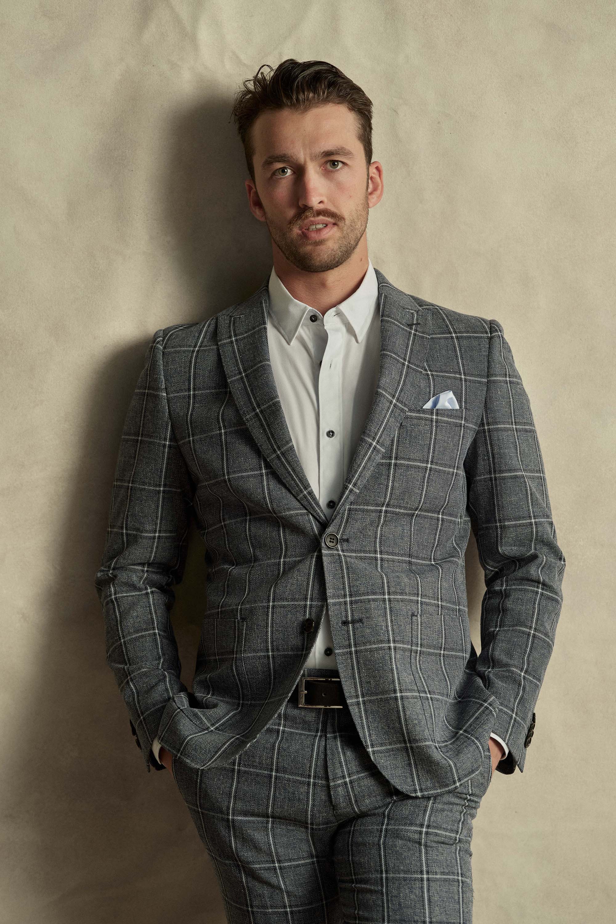 A man stands against a light beige wall wearing a white dress shirt and Barkers Highland Check Suit Pant, hands in pockets. He has short hair, light facial hair, and a crisp pocket square, creating a refined look.