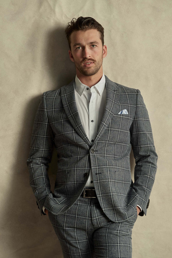 A man stands against a light beige wall wearing a white dress shirt and Barkers Highland Check Suit Pant, hands in pockets. He has short hair, light facial hair, and a crisp pocket square, creating a refined look.