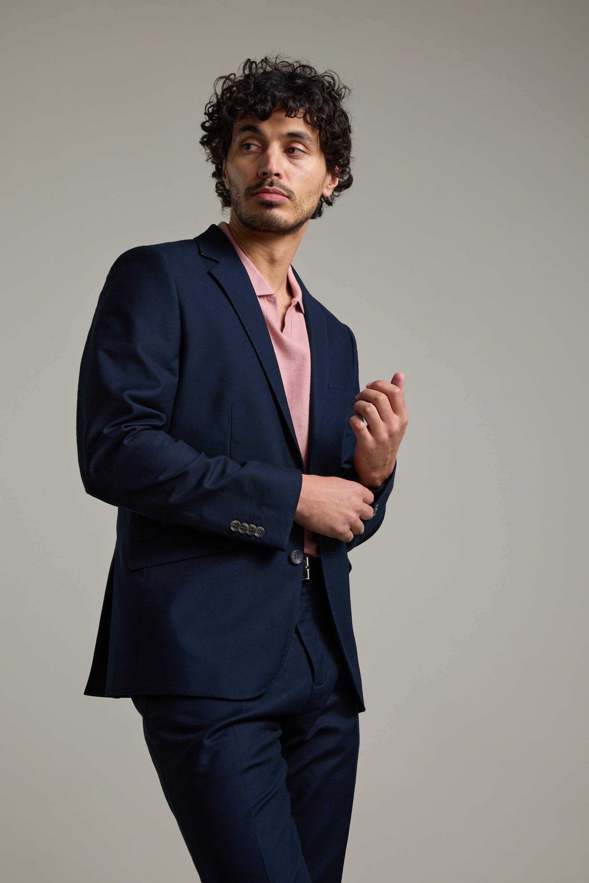 A man with curly hair models the Barkers Moris Cotton Blazer over a light pink shirt, standing against a plain background and gazing thoughtfully to his left, with one hand near his face and the other at his waist.