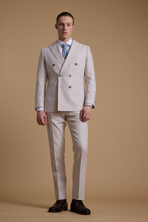 A man wears the Barkers Somerton Linen Double Breasted Blazer with matching trousers, a white shirt, blue patterned tie, and dark brown shoes against a beige background—an elegant summer suit look.
