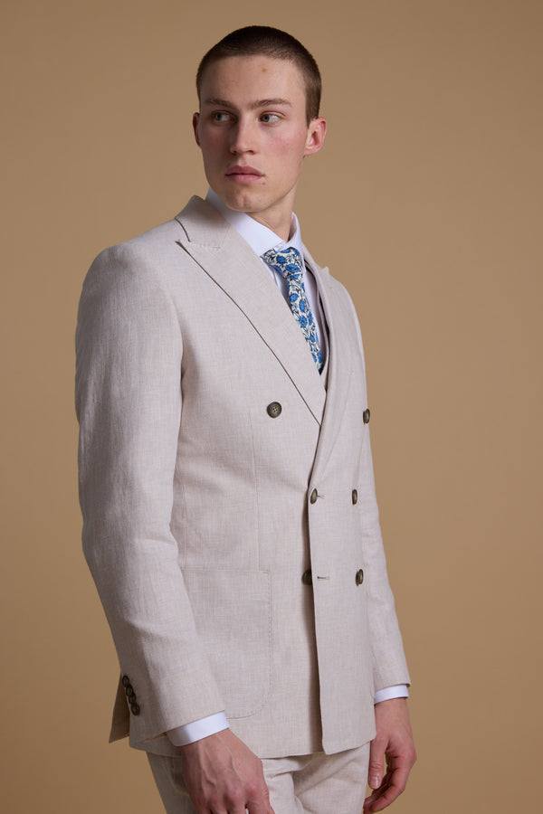 A young man with short hair models the Barkers Somerton Linen Double Breasted Blazer in light beige over a white shirt and blue patterned tie, standing against a plain beige background and looking to his left with a neutral expression.