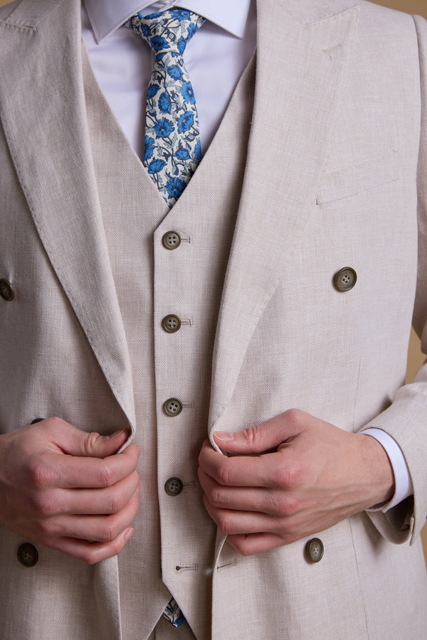 A person adjusts their Somerton Linen Double Breasted Blazer by Barkers, featuring brown buttons and a blue floral tie. The cropped shot from neck to waist highlights the blazer's details and the wearer’s hands.