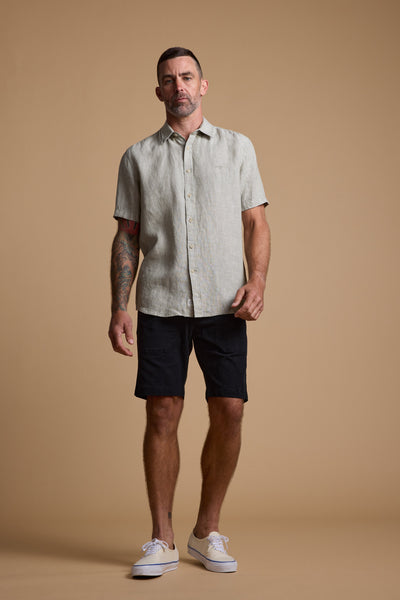 A man with short hair, a beard, and tattoos on his left arm stands against a tan background wearing Barkers' Tidal GD Utility Short in black, a garment-dyed beige button-up shirt, and white sneakers.