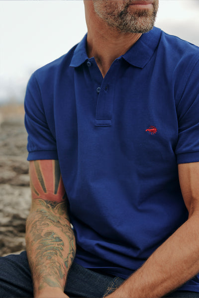 A man with a short beard and tattooed arms wears a blue Barkers Regent Pique Polo, made from organic cotton with a small red crab logo, as he sits outdoors on rocky ground.